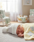 Stork And Baby white knit infant earflap hat with pom-pom, on sleeping baby with yellow elephant blanket.