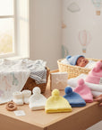 Stork And Baby pastel knit earflap hats in pink, yellow, and blue with pom-poms. A baby sleeps in the background.