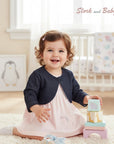 Stork and Baby dark denim bolero worn by a smiling infant girl in a pink unicorn dress, sitting on a rug.