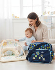 Stork And Baby navy nappy bag with fun animal print, multiple compartments, and adjustable strap. A mother holds her baby on a play mat.