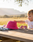 Stork And Baby pink cotton bucket hat with a white and blue elephant print swaddle and pink sandals on a wooden table with a baby reaching for the hat.