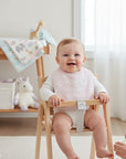 Stork And Baby pink patterned muslin bib on a smiling baby sitting in a high chair. Soft, breathable fabric for feeding.