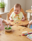 Three children sitting at a wooden table with food and toys, in a home setting.