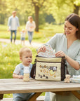 Stork And Baby Small beige diaper bag with bear embroidery, organized compartments, and a checkered trim. A woman places items inside while a baby sits nearby.
