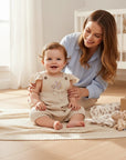 Stork And Baby beige muslin romper with flutter sleeves and bunny embroidery on a baby sitting on a blanket.
