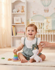 Stork And Baby girl in a blue cotton linen 2-piece set with white eyelet trim and a floral embellishment, playing with toys.