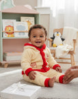 Stork And Baby girls' cream coral fleece tracksuit with red trim and "Too Cute" embroidery. Baby sitting on floor.