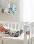 Stork And Baby grey infant socks with white lace trim and large grey bows with pearl centers. A baby's feet are visible in a crib.