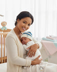 Stork And Baby striped knit infants winter hat with a penguin logo, worn by a sleeping baby held by a mother.