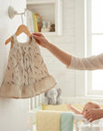 Stork and Baby natural muslin dress with lavender print and ruffled hem. A mother holds it up in a nursery.