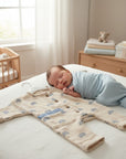 Stork And Baby beige muslin romper with blue rainbow print and button closure, on a bed next to a sleeping baby.