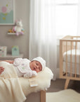 Stork And Baby newborn baby girl in a white floral print outfit and hat sleeping on a fluffy rug next to a crib.