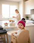 Stork And Baby infant winter hat with pink, blue, and yellow stripes and a navy pom-pom. A smiling baby sits in a high chair.