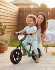 Stork And Baby green toddler balance bike with a smiling child riding and a woman kneeling behind them.