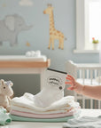 Stork And Baby white cotton infant socks with a textured pattern, size 0-6 months, on a stack of baby clothes.